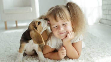 Puppy licking little girls face while they sit together on a white carpet