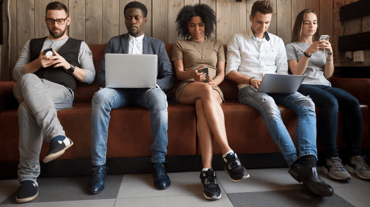 5 millennials sitting on a bench on various electronic devices such as smartphone and laptops